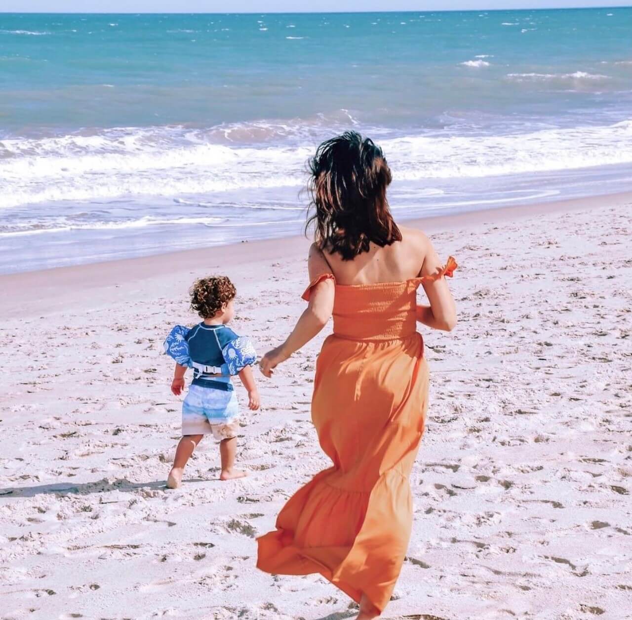 A woman in an orange dress and a young child walking toward the ocean on a sandy beach in Vero Beach, FL