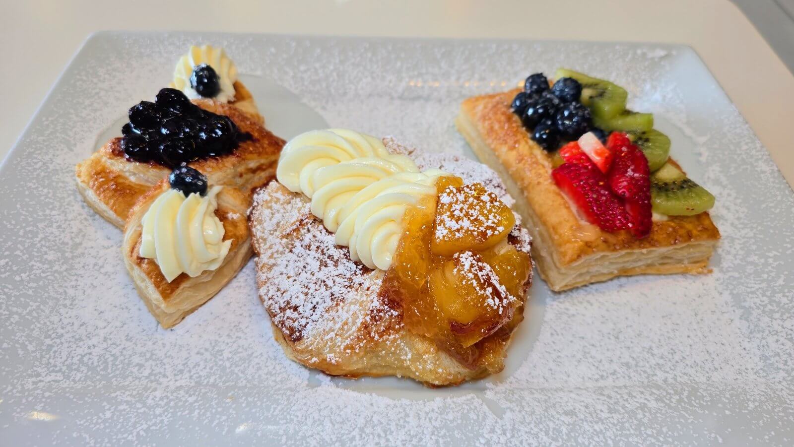 A plate of assorted fruit-topped pastries dusted with powdered sugar, including one with blueberries and cream, one with caramelized peaches and cream, and one with strawberries, kiwi, and blueberries.