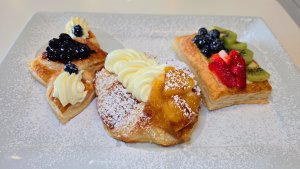 A plate of assorted fruit-topped pastries dusted with powdered sugar, including one with blueberries and cream, one with caramelized peaches and cream, and one with strawberries, kiwi, and blueberries.
