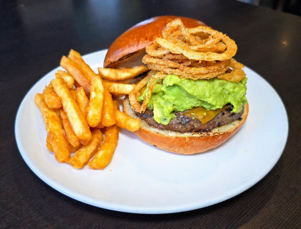 Dinner Revolutions Guac Burger with the top bun slanted off showing the large serving of guac on the burger as well as some crispy fried onion straws. Plated with fires.