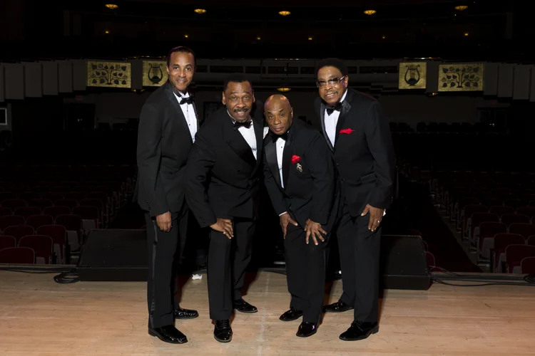 The band, The Drifters, in matching black tuxedos and bow ties pose together on an empty stage